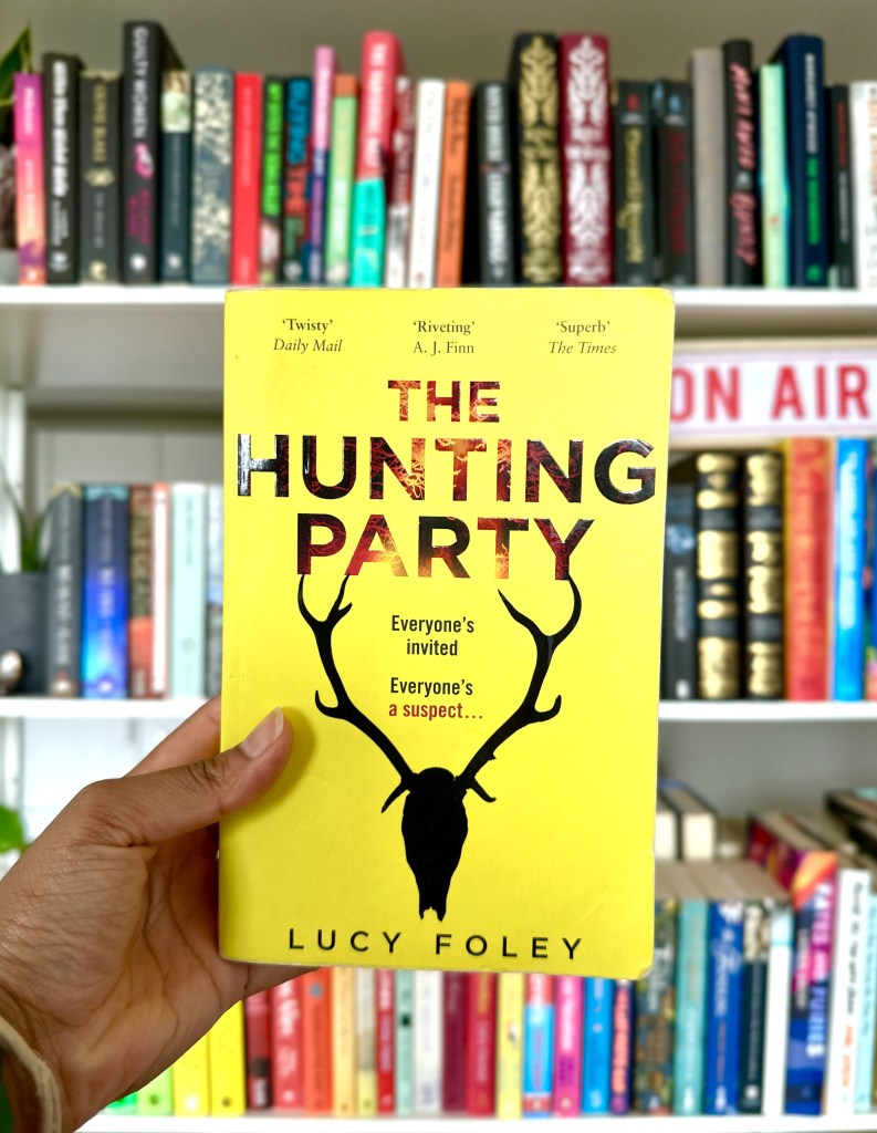 Photo of The Hunting Party book by Lucy Foley in front of a bookshelf. Yellow book cover with black silhouette of a deer.